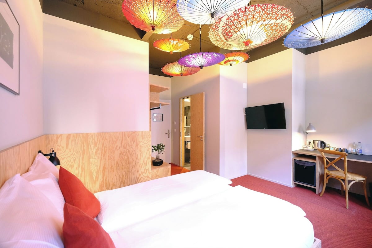 A hotel room with a bed, a desk, a TV, and ceiling lamps.