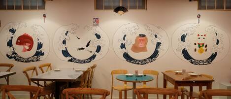 Un restaurante con mesas y sillas de madera, una pared blanca con motivos de olas y un abanico japonés tradicional.