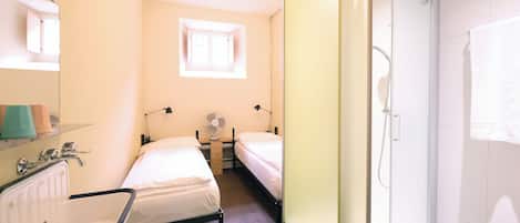 Une chambre d’hôtel de taille réduite, comprenant un lit simple, une petite table de chevet, un miroir, un lavabo et un espace douche.