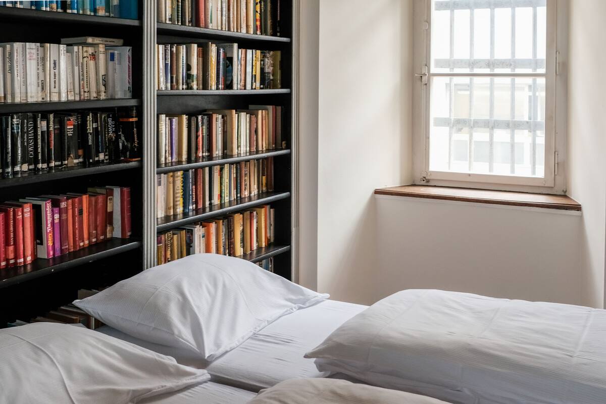 A room with a large bookshelf filled with books, a window with bars, and two beds with white bedding.