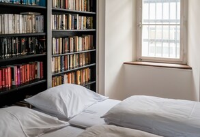 Una habitación con una amplia estantería llena de libros, una ventana con rejas y dos camas con ropa blanca.