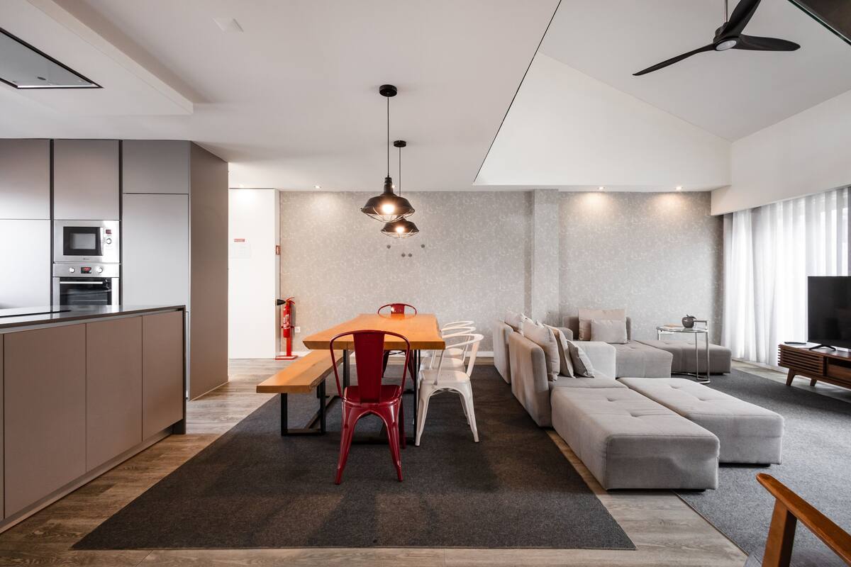 A modern kitchen with built-in appliances, a dining area with a wooden table and chairs, a grey sofa, and a ceiling fan.