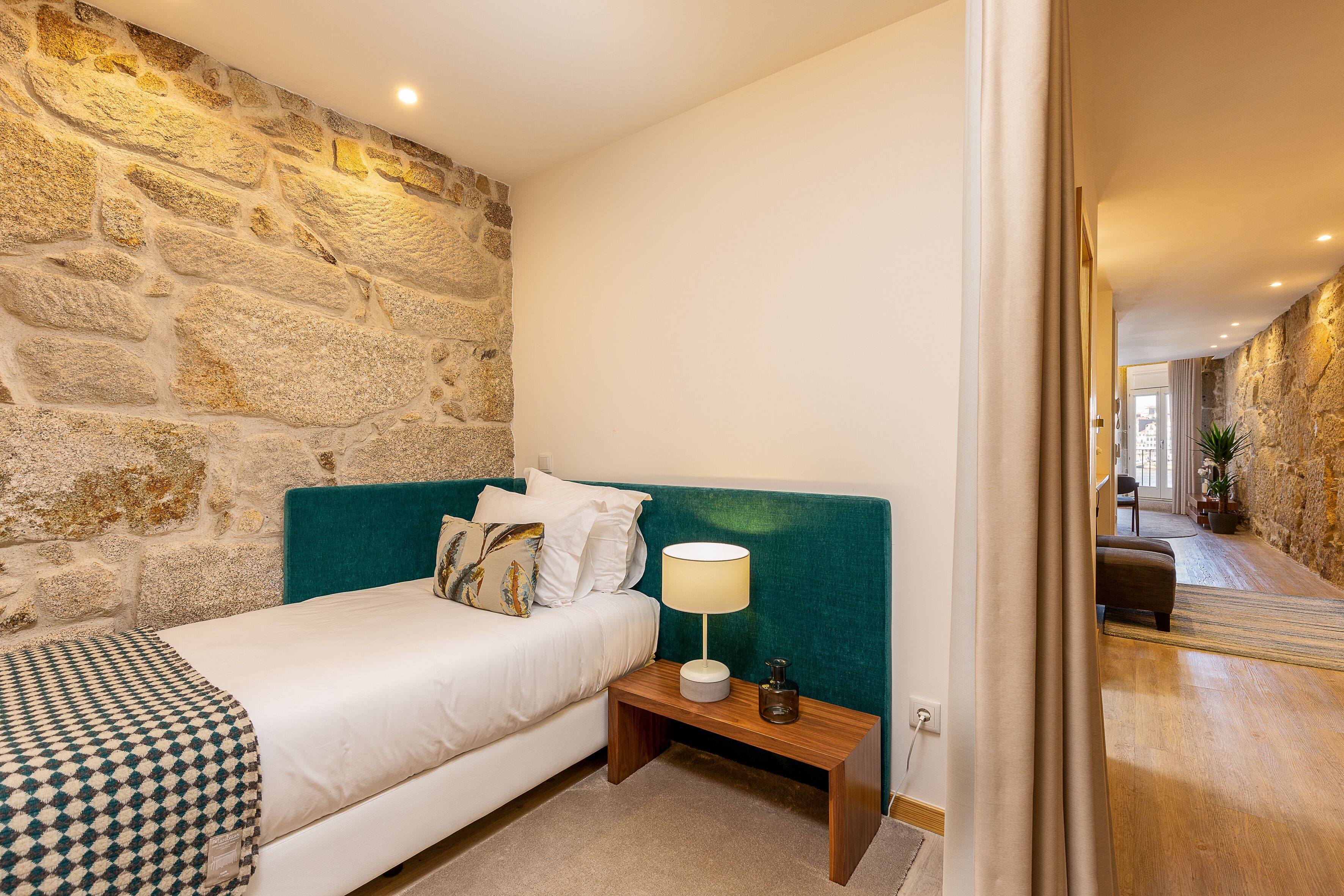 Une chambre avec un mur en pierre, une tête de lit de couleur turquoise, un lit recouvert d’une couverture à carreaux, une table de chevet avec une lampe et une petite table de nuit avec une bouteille.