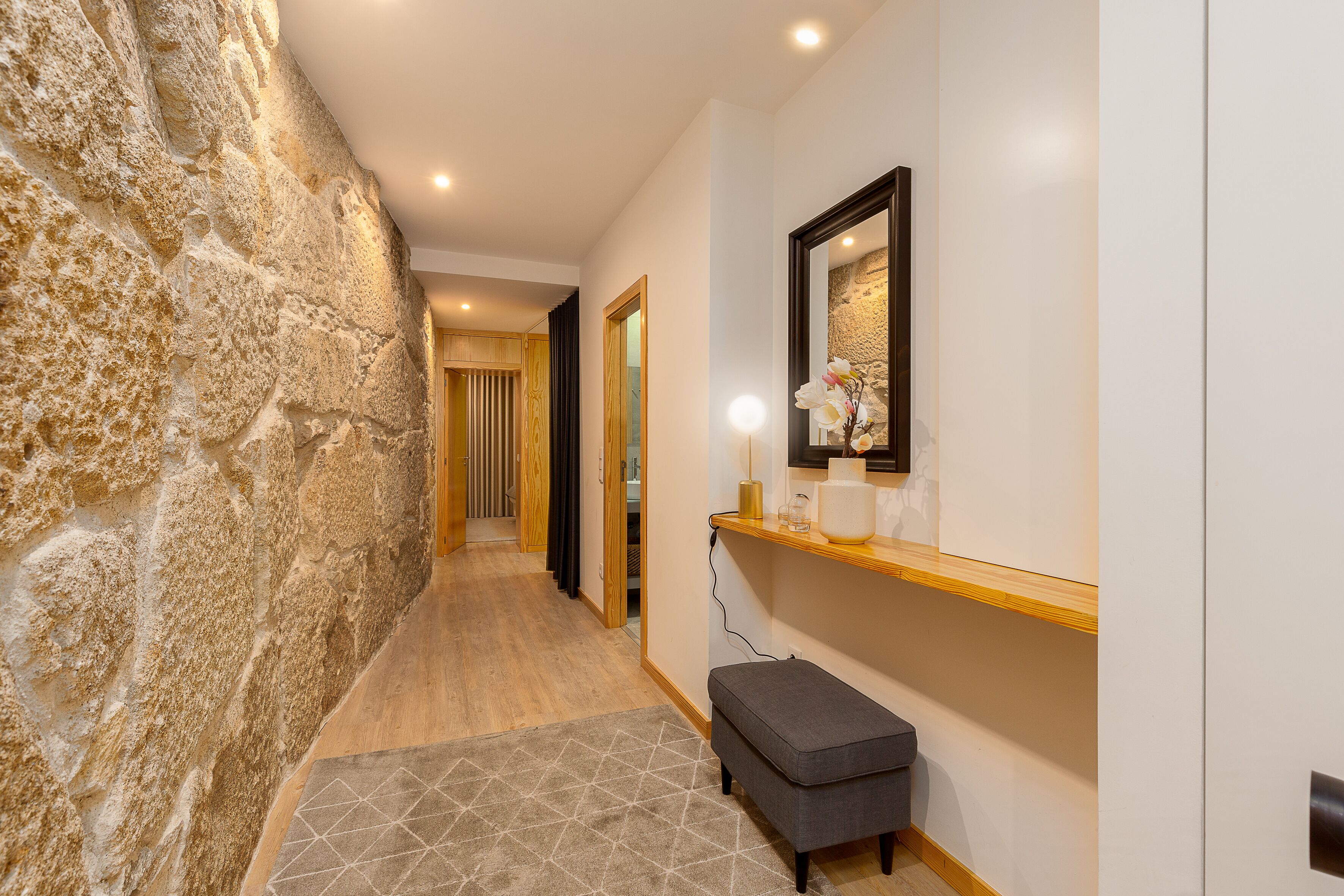 Un pasillo con pared de piedra, un banco de madera, un espejo y una estantería con un jarrón.