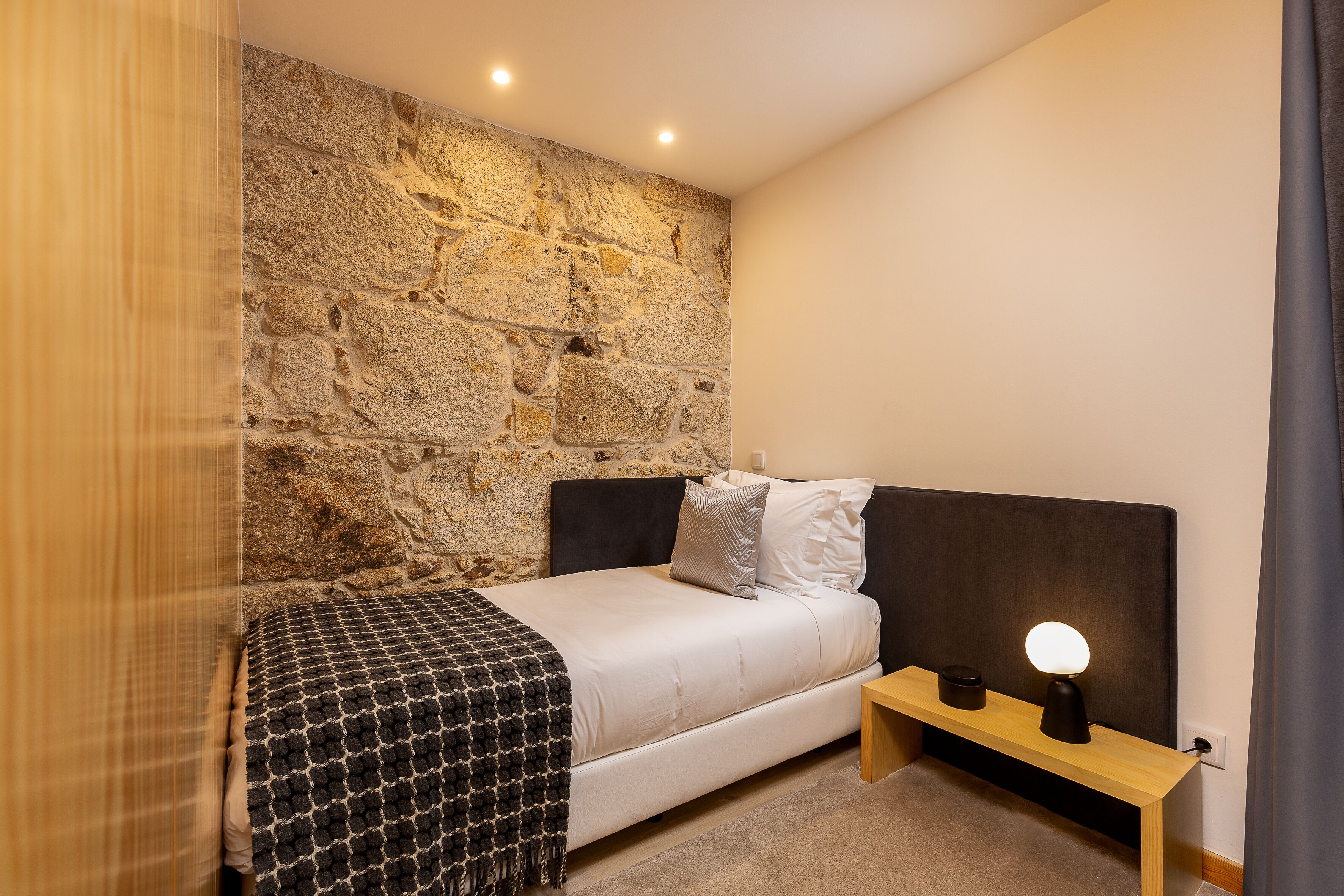 A bedroom with a stone wall, a bed with a headboard, a bedside table with a lamp, and a window with curtains.