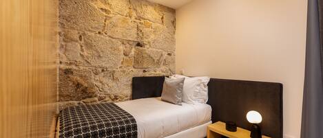 A bedroom with a stone wall, a bed with a headboard, a bedside table with a lamp, and a window with curtains.