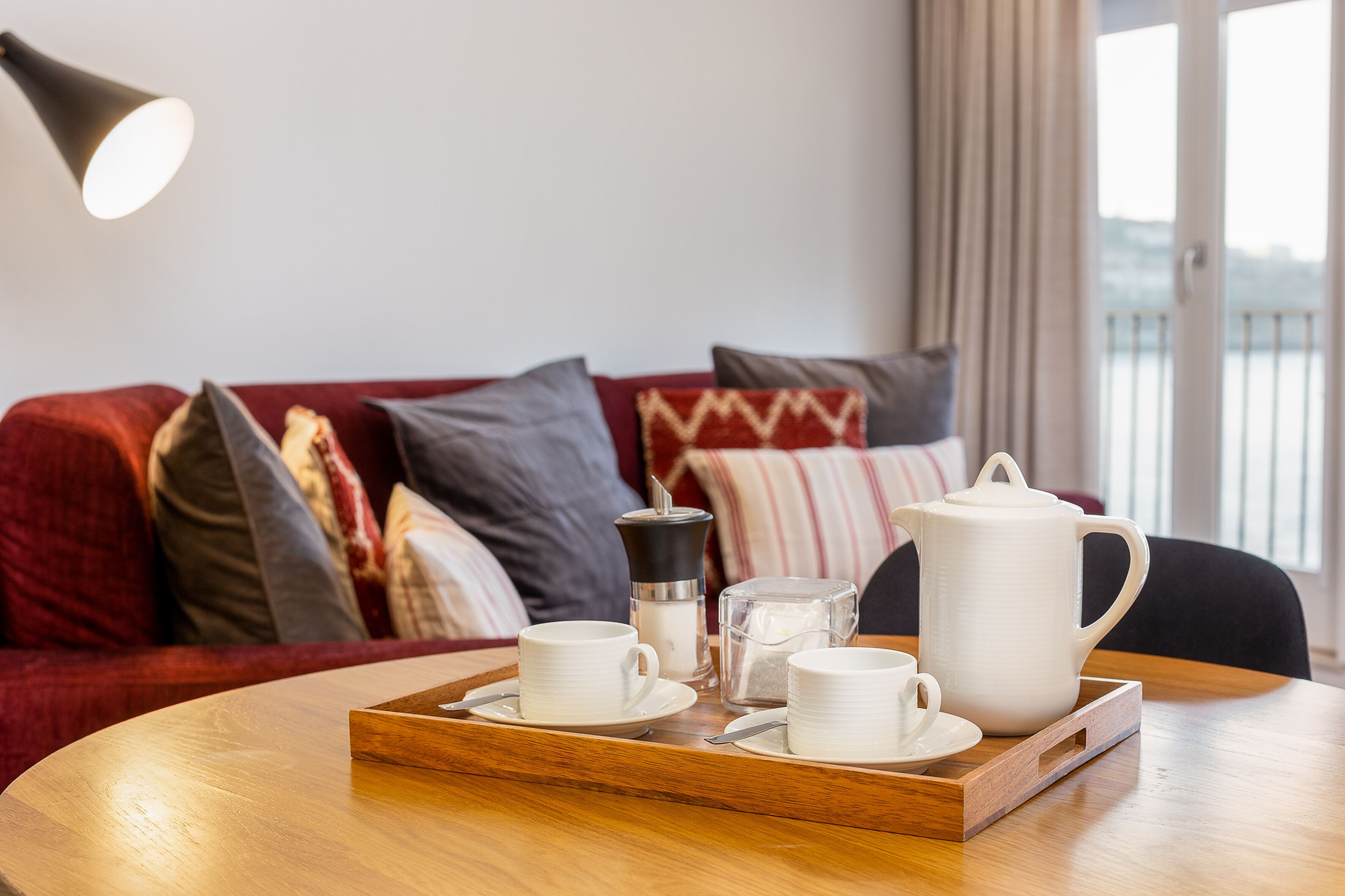 Una bandeja de madera con tazas blancas, un hervidor de agua y un recipiente de vidrio sobre una mesa redonda en una habitación con un sofá rojo y vistas a un balcón.