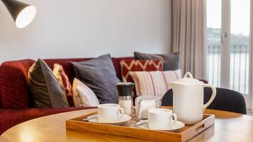 Una bandeja de madera con tazas blancas, un hervidor de agua y un recipiente de vidrio sobre una mesa redonda en una habitación con un sofá rojo y vistas a un balcón.