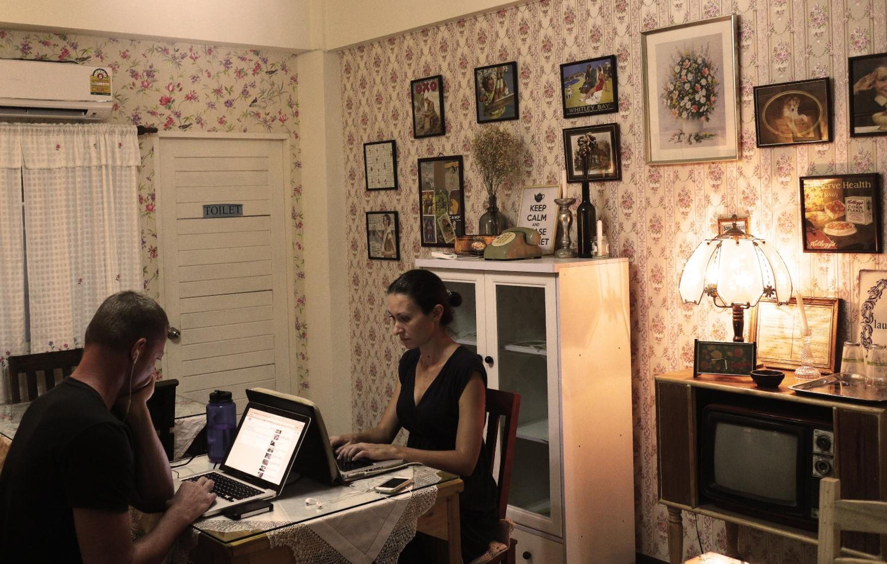 A room with a white door labeled 'TOILET', a table with a laptop, a person using a laptop, a microwave, and a cabinet with various items.
