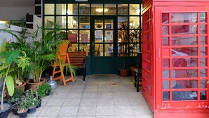A red telephone booth labeled "TEA ROOM" with a green door and windows, surrounded by potted plants and a wooden chair. - The London Tearoom (Chiang Mai)