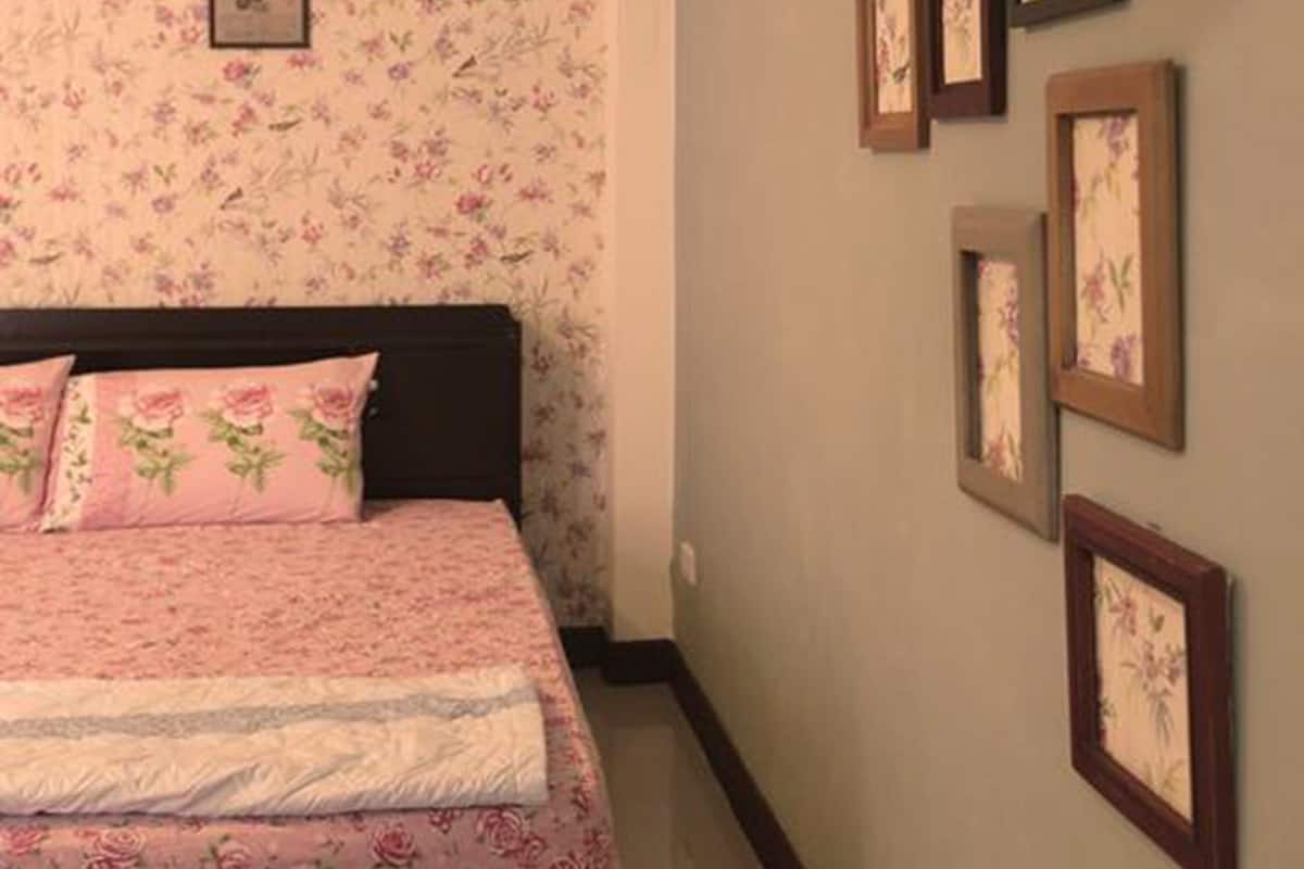 A bedroom with a floral wallpaper, a bed with pink bedding, and several framed pictures on the wall.