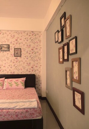 A bedroom with a floral wallpaper, a bed with pink bedding, and several framed pictures on the wall.