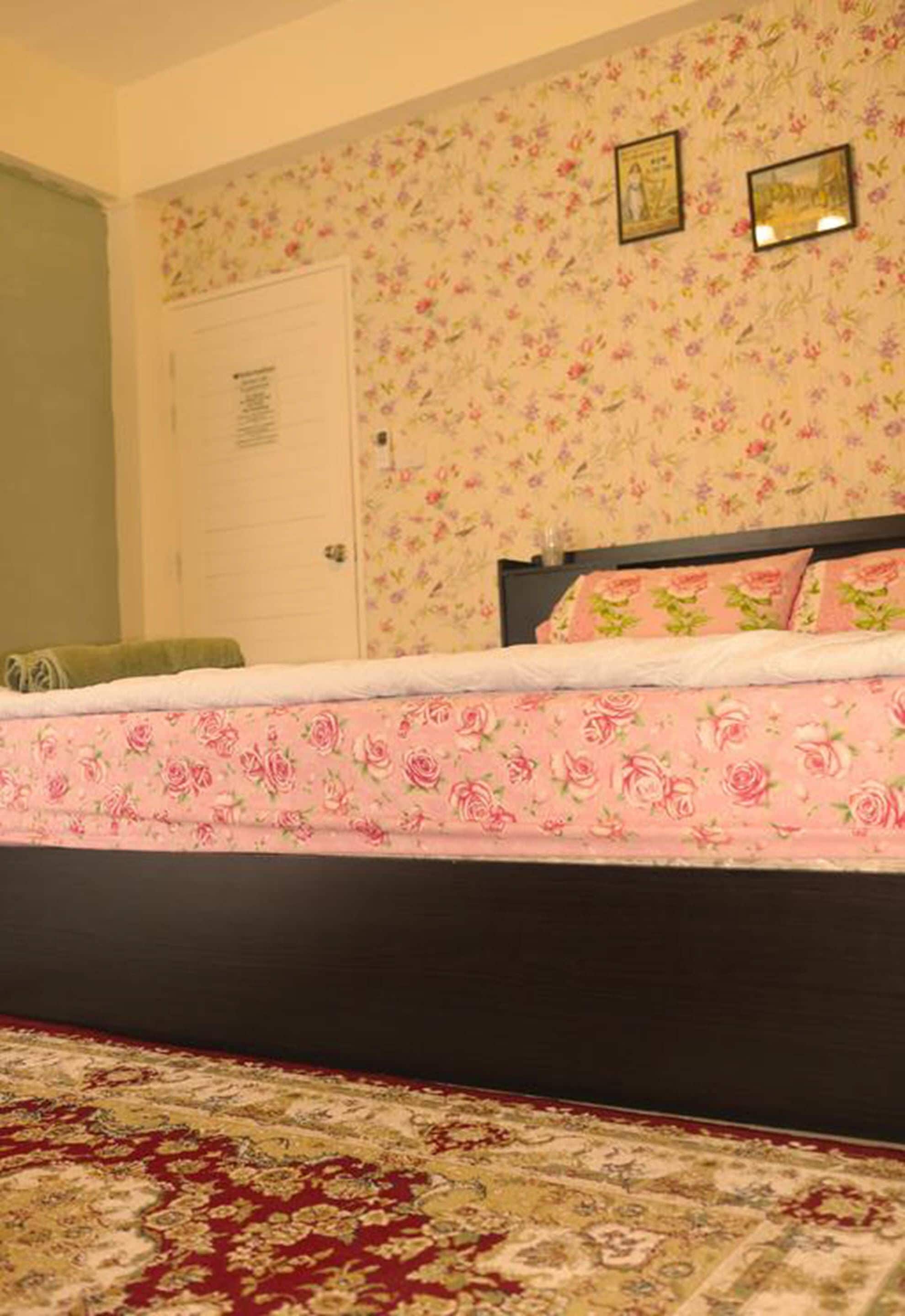 Quarto com papel de parede floral, cama com colcha floral rosa e tapete vermelho estampado.