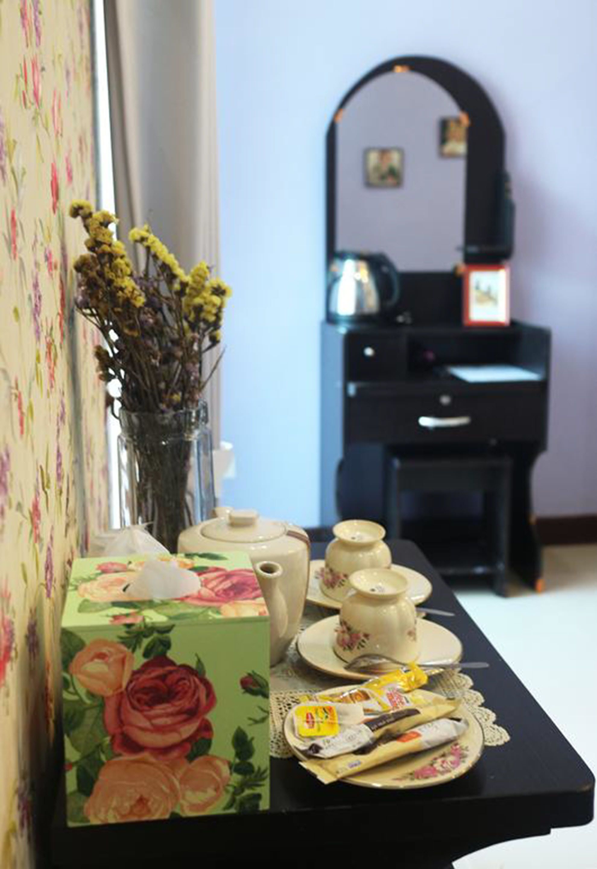 A room with floral wallpaper, a vase with dried flowers, a tea set, and a desk with a computer and a mirror.