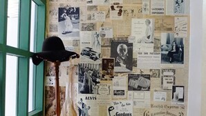 A room with a wall covered in vintage posters and clippings, a vintage hat on a stand, a small table with a vase, and a tufted armchair. - The London Tearoom (Chiang Mai)