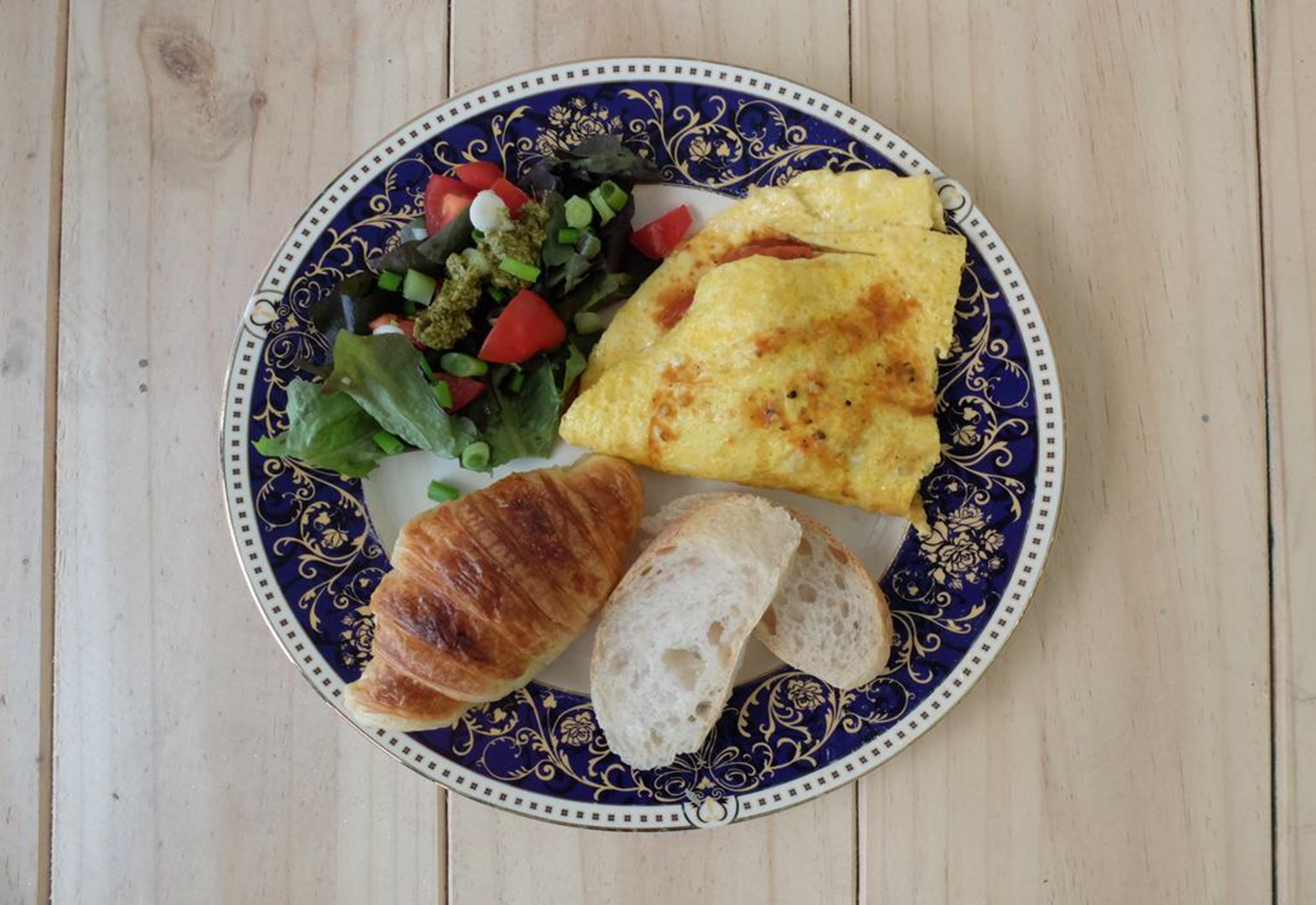 A plate with a croissant, bread, omelette, and salad on a decorative plate.