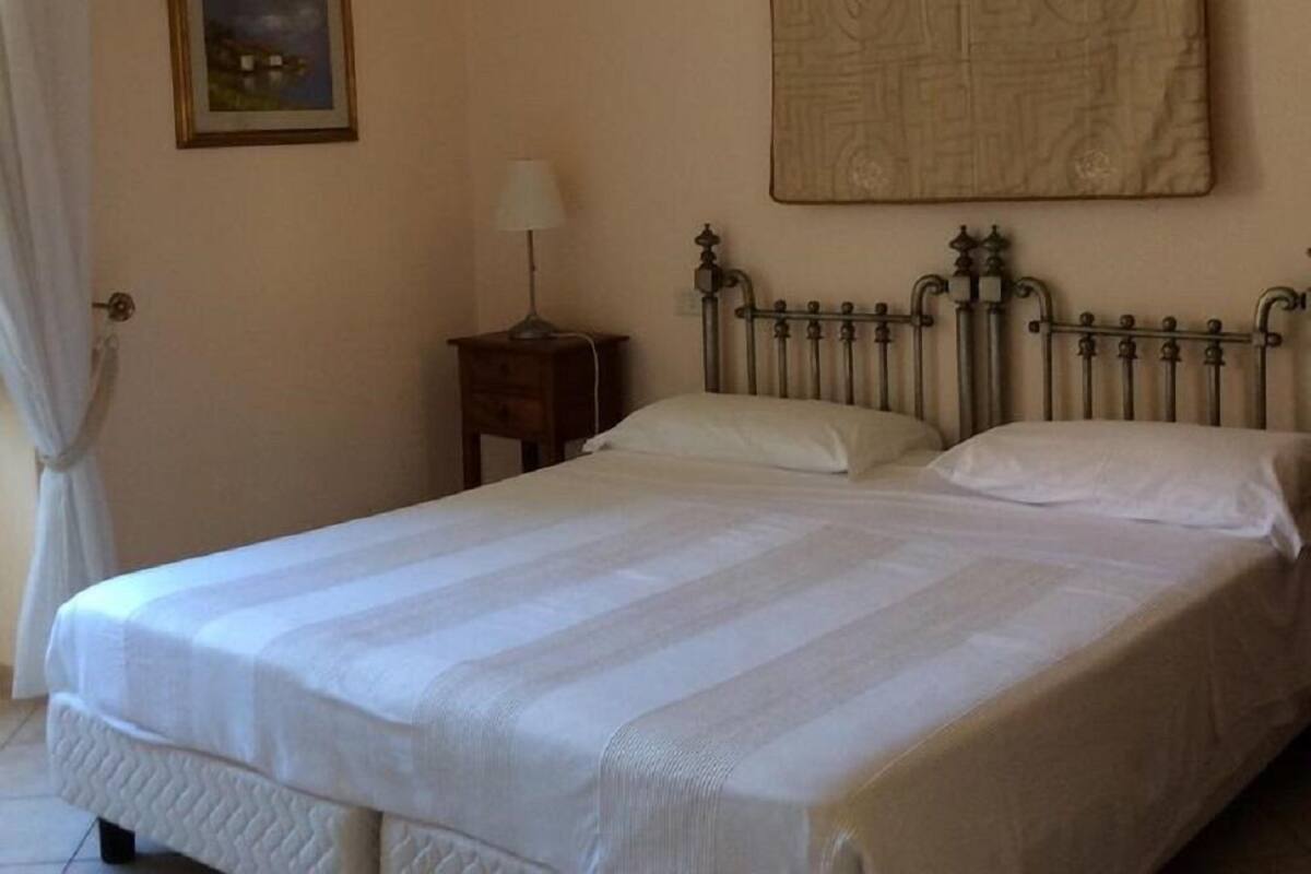 Un lit bien fait, avec une tête de lit, une table de chevet avec une lampe, un tableau encadré au mur et une fenêtre avec des rideaux.
