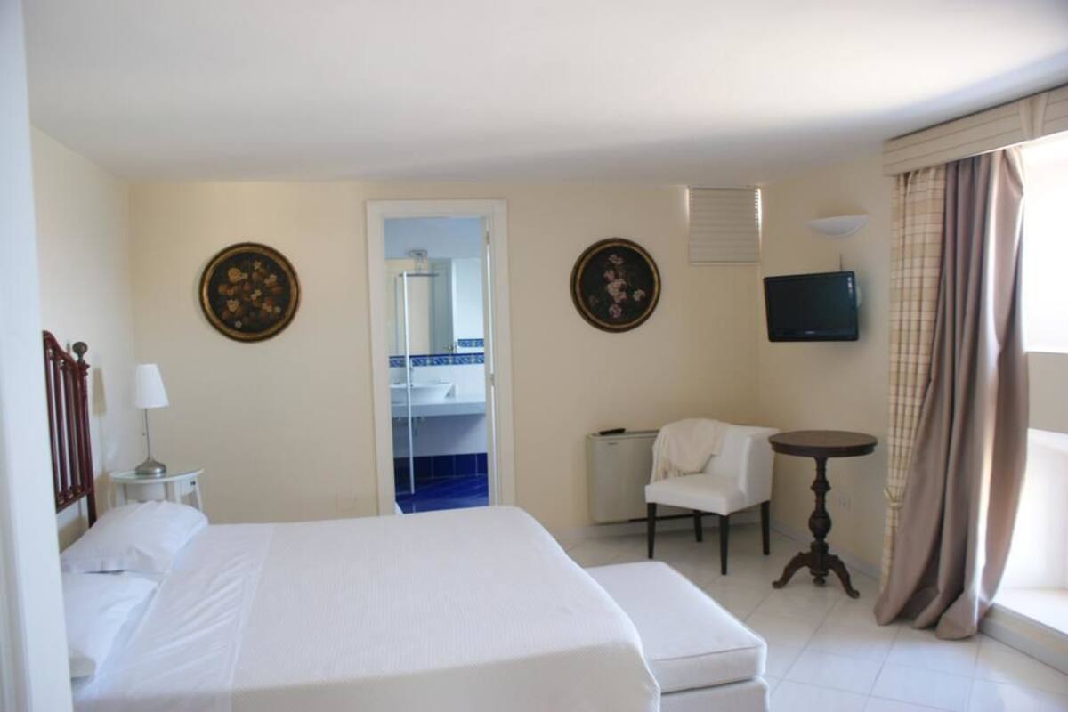 Une chambre d’hôtel avec un grand lit, une petite table, une chaise, un téléviseur et une salle de bain visible à travers une porte.