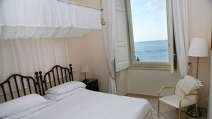 A bedroom with a bed, a chair, a nightstand, a lamp, and a view of the sea through an open window. - Palazzetto Florio (Castellabate)