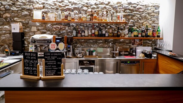 Zona de bar con estantes llenos de botellas de licor, una máquina de café y un menú en pizarra que enumera diversas bebidas.