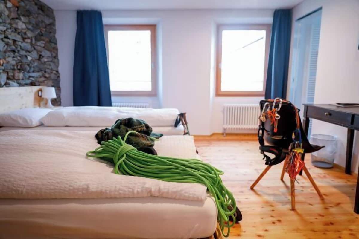 Une chambre d’hôtel avec un lit, un mur en pierre, deux fenêtres avec des rideaux bleus et un bureau avec une chaise.