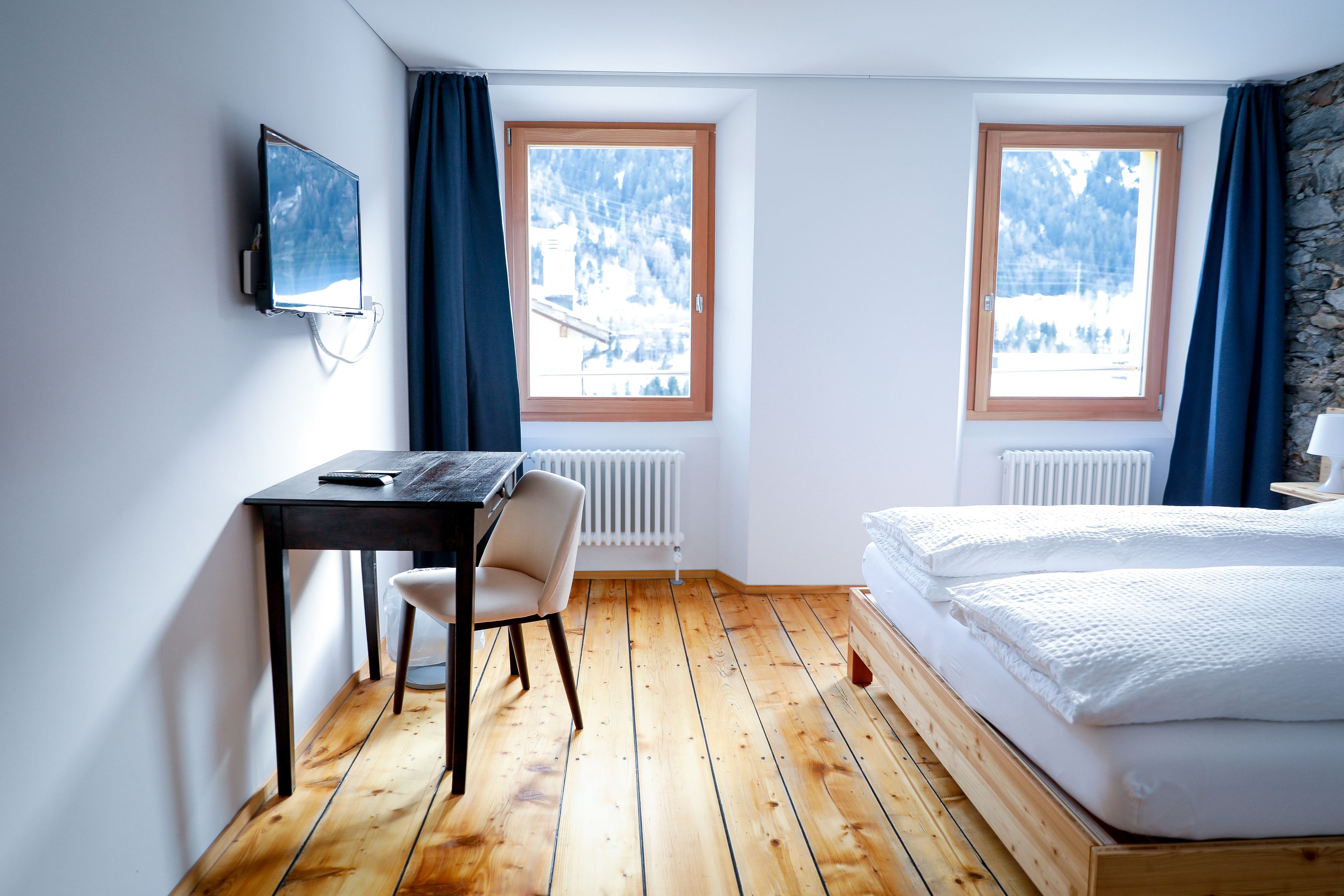 A room with a wooden floor, a bed with white bedding, a desk with a chair, and two windows with curtains.