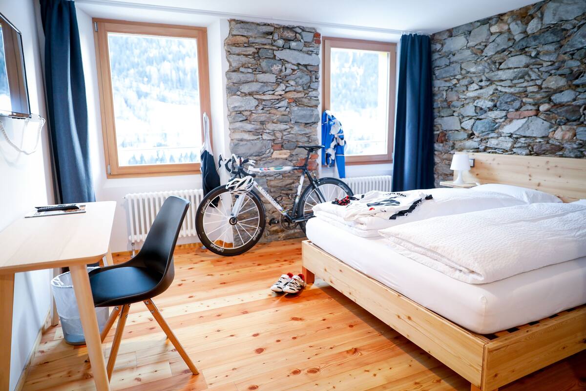 Une chambre avec un lit en bois, un bureau, une chaise, un vélo et un mur en pierre.