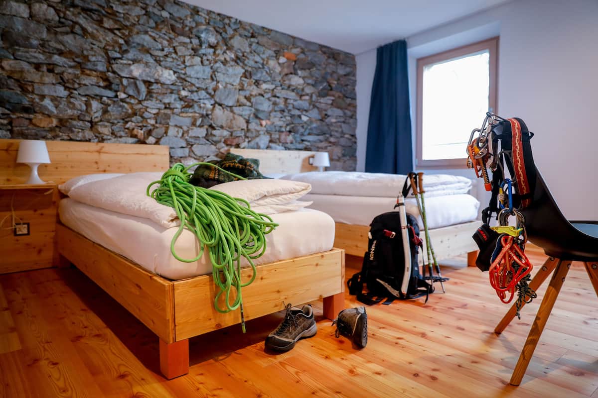 Une chambre avec un mur en pierre, un lit en bois, deux oreillers, une corde verte, un sac à dos et une chaise avec du matériel d’escalade.