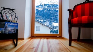 Una habitación con piso de madera, una silla roja, una estantería con libros y vista a un paisaje nevado de montaña a través de una ventana.