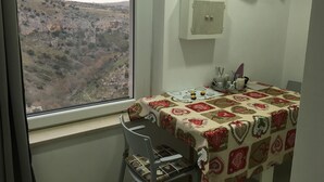 Una habitación con un ventanal grande que ofrece vistas a un paisaje montañoso. Hay una mesa con mantel estampado, una silla y un pequeño aparador con objetos decorativos.