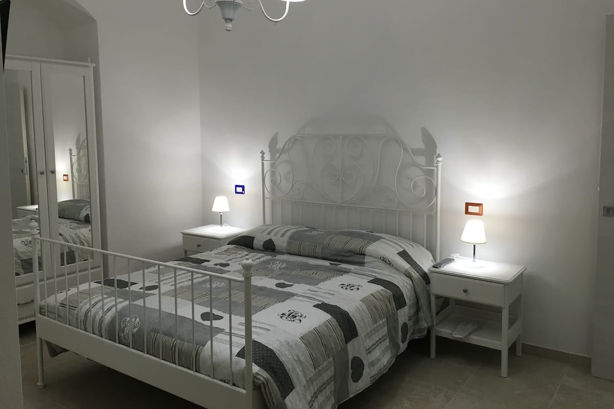 A bedroom with a bed, two bedside tables, a nightstand, a mirror, and a chandelier.