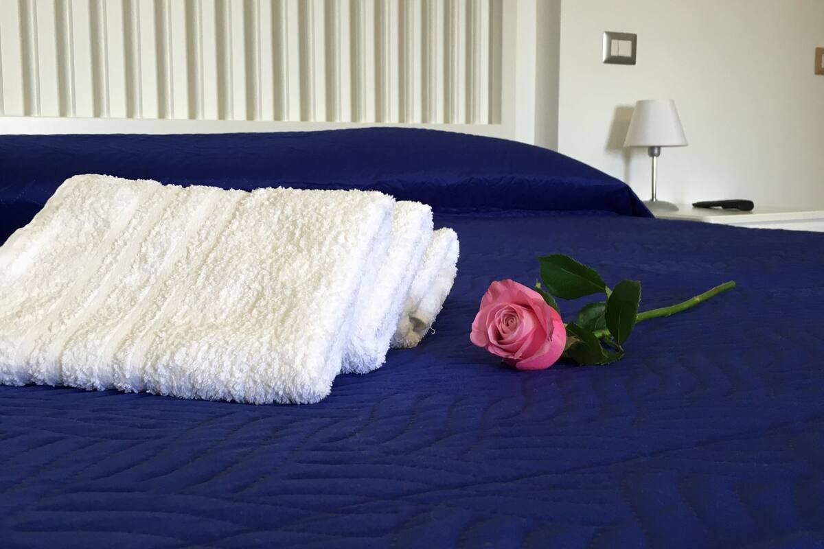 A neatly made bed with a folded white towel and a pink rose on a dark blue bedspread.