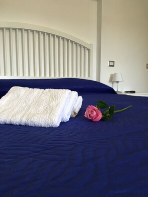A neatly made bed with a folded white towel and a pink rose on a dark blue bedspread. - Il Peccato Originale (Matera)