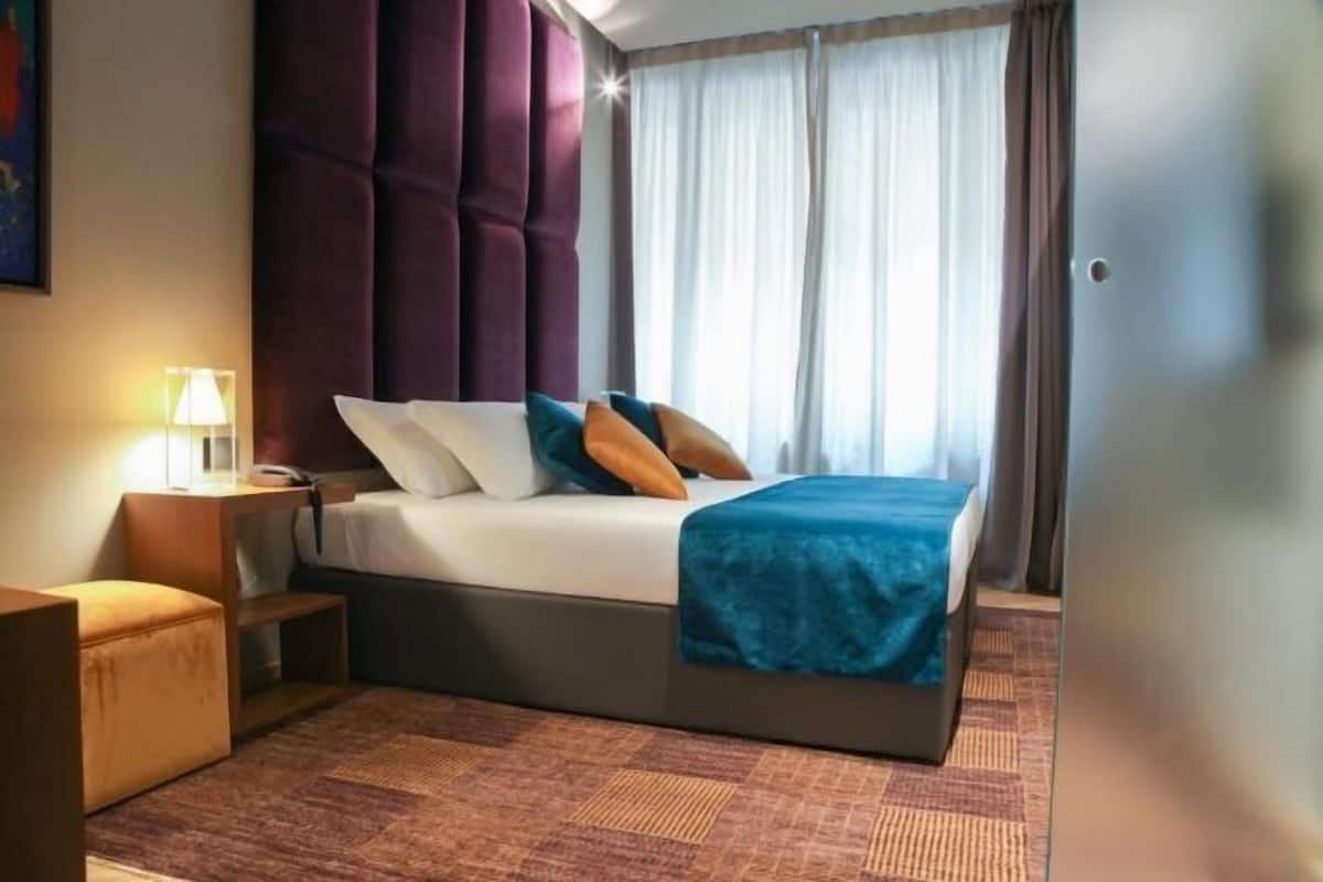 Una camera d'albergo con un letto, un comodino, una lampada e vista all'esterno attraverso la finestra.