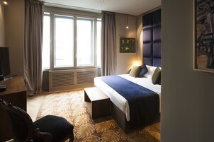 Una camera d'albergo con un letto grande, una scrivania, una sedia, una finestra con tende e un quadro appeso al muro.