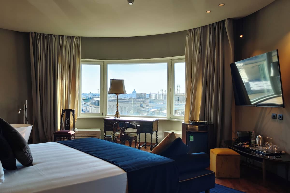 Una camera d'albergo moderna con un letto grande, una scrivania con una lampada, una TV e vista sulla città.