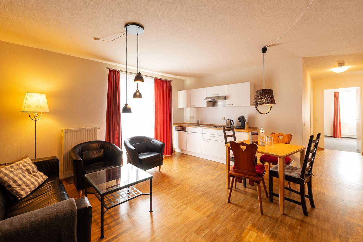 A modern living room with a sofa, a dining table with chairs, a kitchen area, and a red curtain.