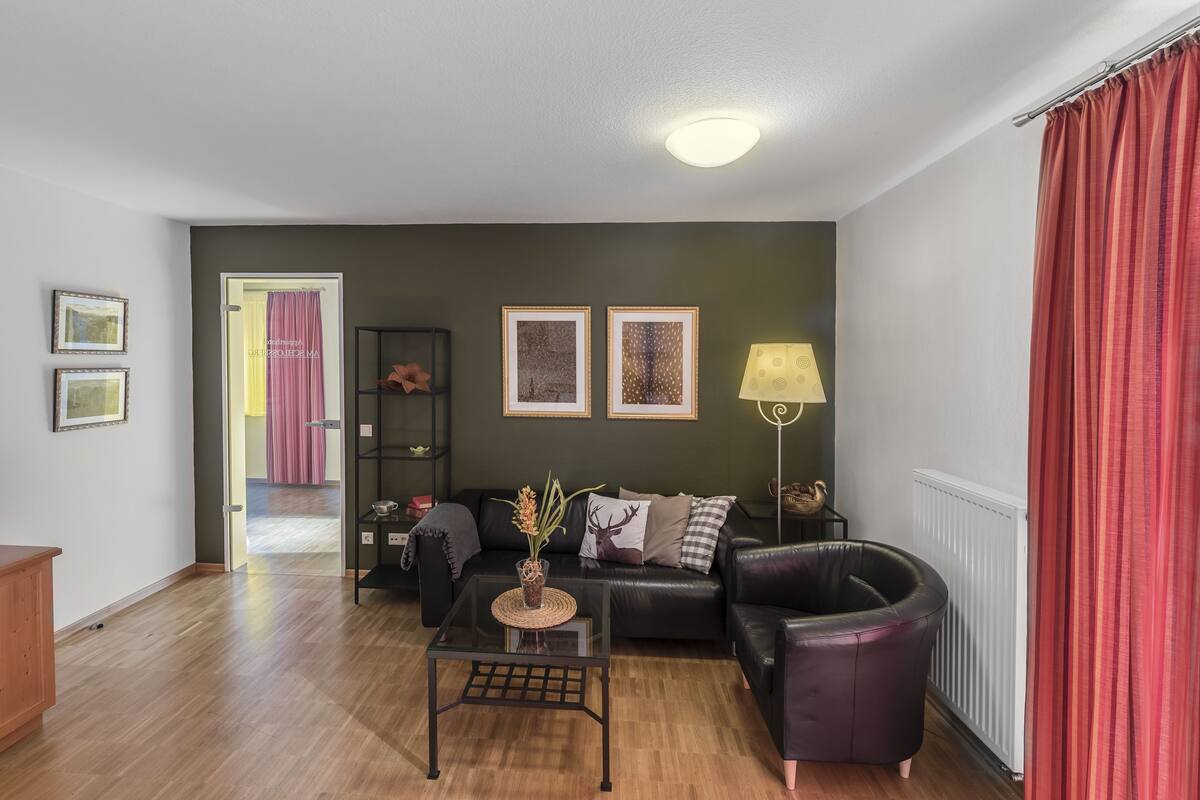 A modern living room with a black leather sofa, a glass coffee table, and a lamp. There are framed pictures on the wall and a door leading to another room.