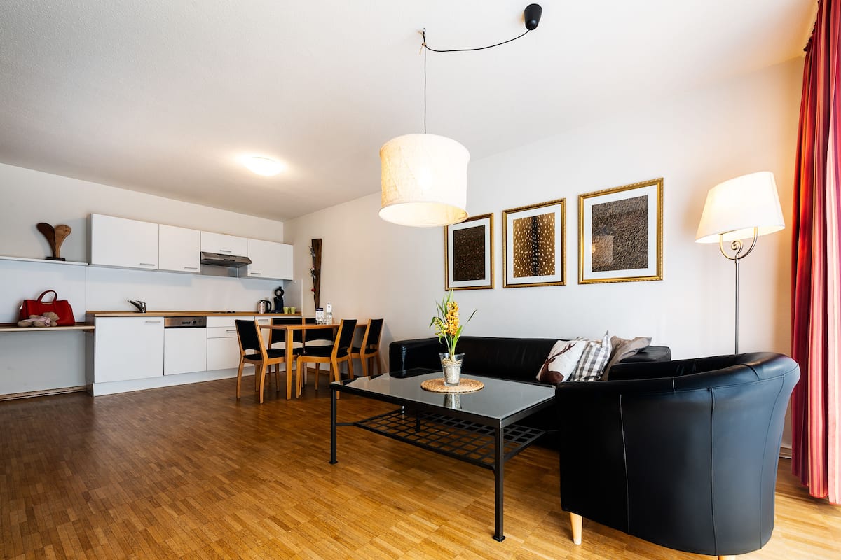 A modern living room with a black leather sofa, a glass coffee table, and a dining area with wooden chairs and a white table. There are framed artworks on the wall and a red curtain on the right.