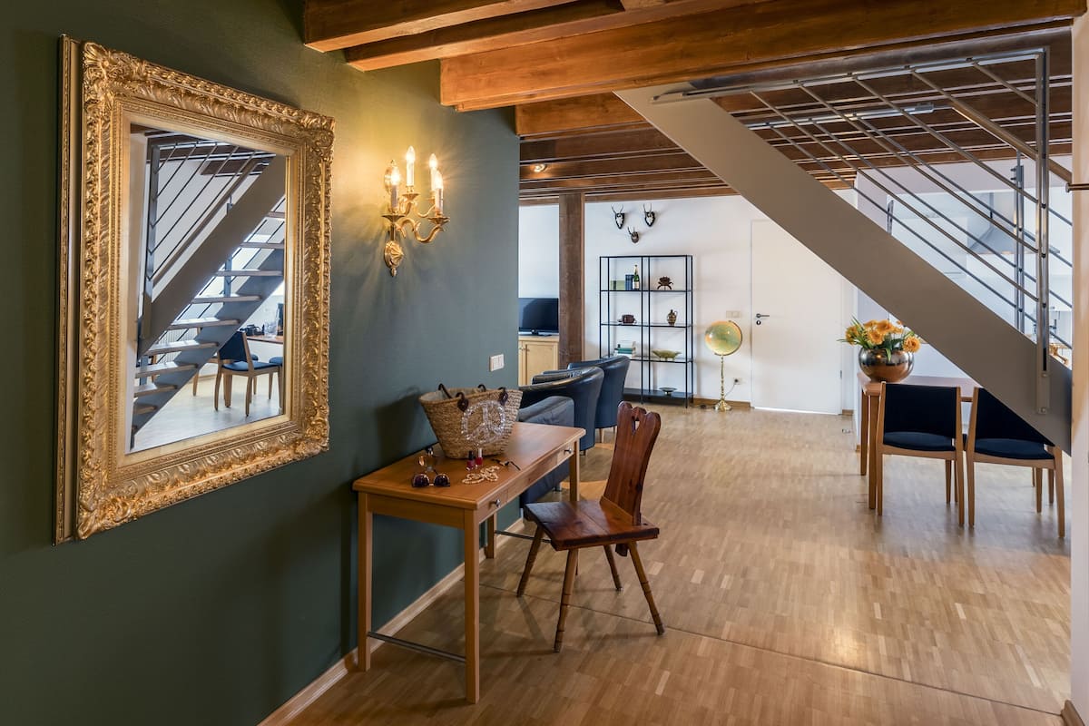 A spacious living area with a staircase, a dining table with chairs, a large mirror, and a bookshelf.