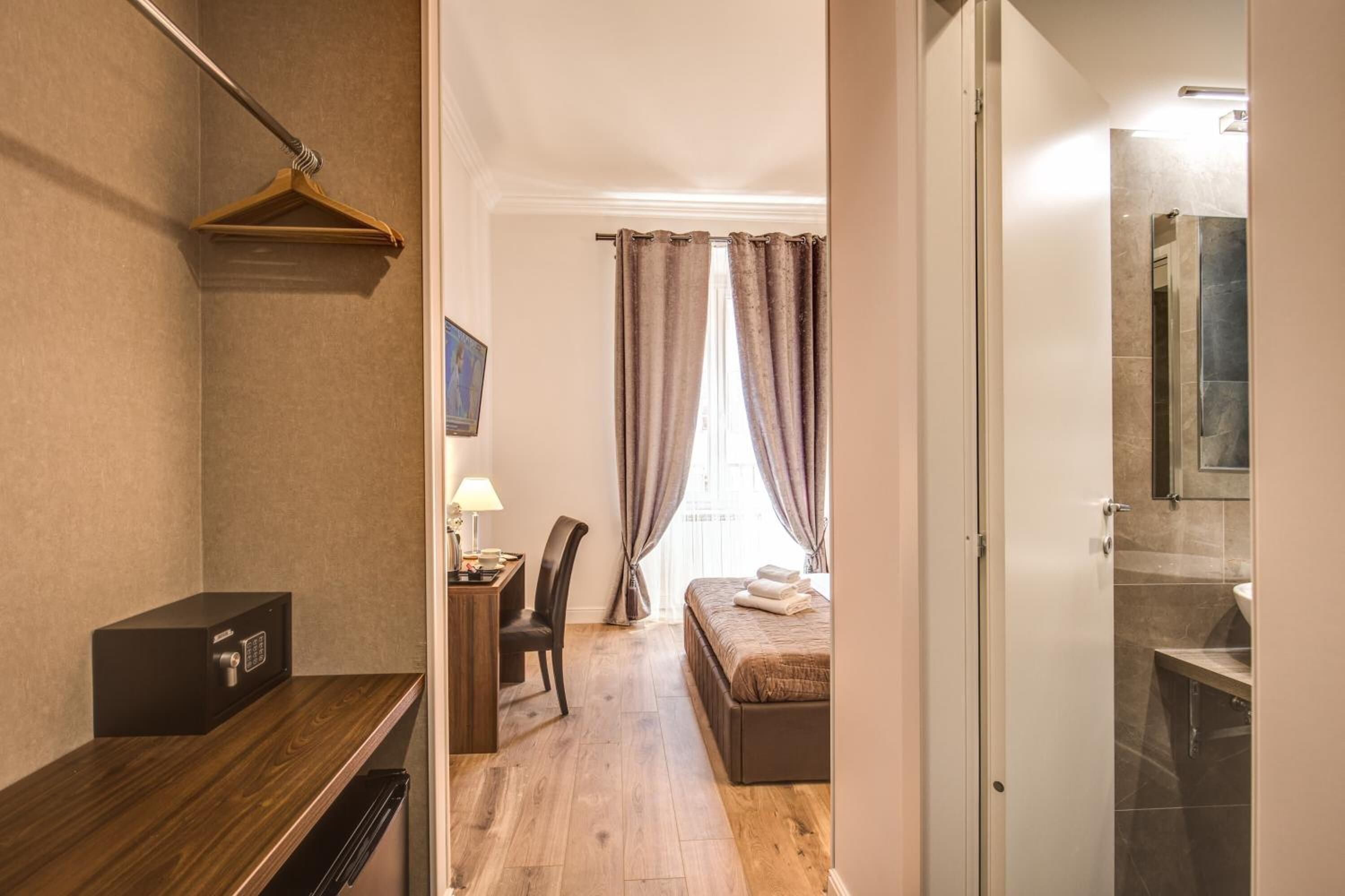 A hotel room with a wooden desk, a bed with a headboard, a chair, a desk lamp, and a window with curtains.