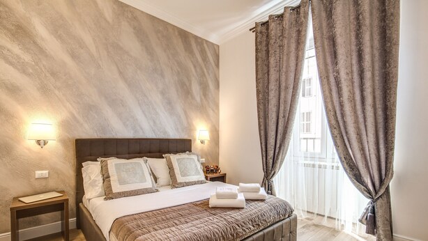 A bedroom with a large bed, two bedside lamps, a nightstand, and a window with curtains. - River Luxury Suites (Rome)