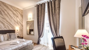 A hotel room with a marble-patterned wall, a bed with a headboard, a bedside table with a lamp, a chair, and a view of the outdoors through a window with curtains. - River Luxury Suites (Rome)