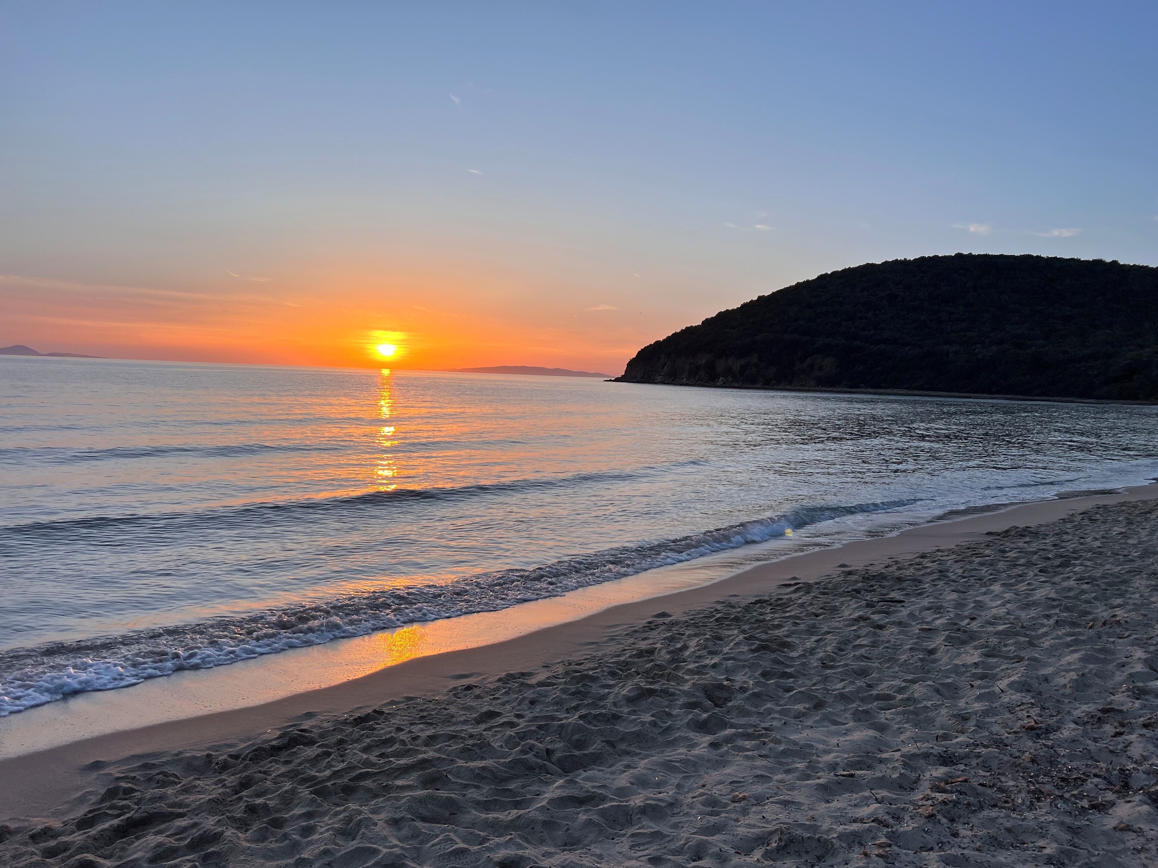 A beach with a sunset over the water, a sandy shore, and a hill in the background.