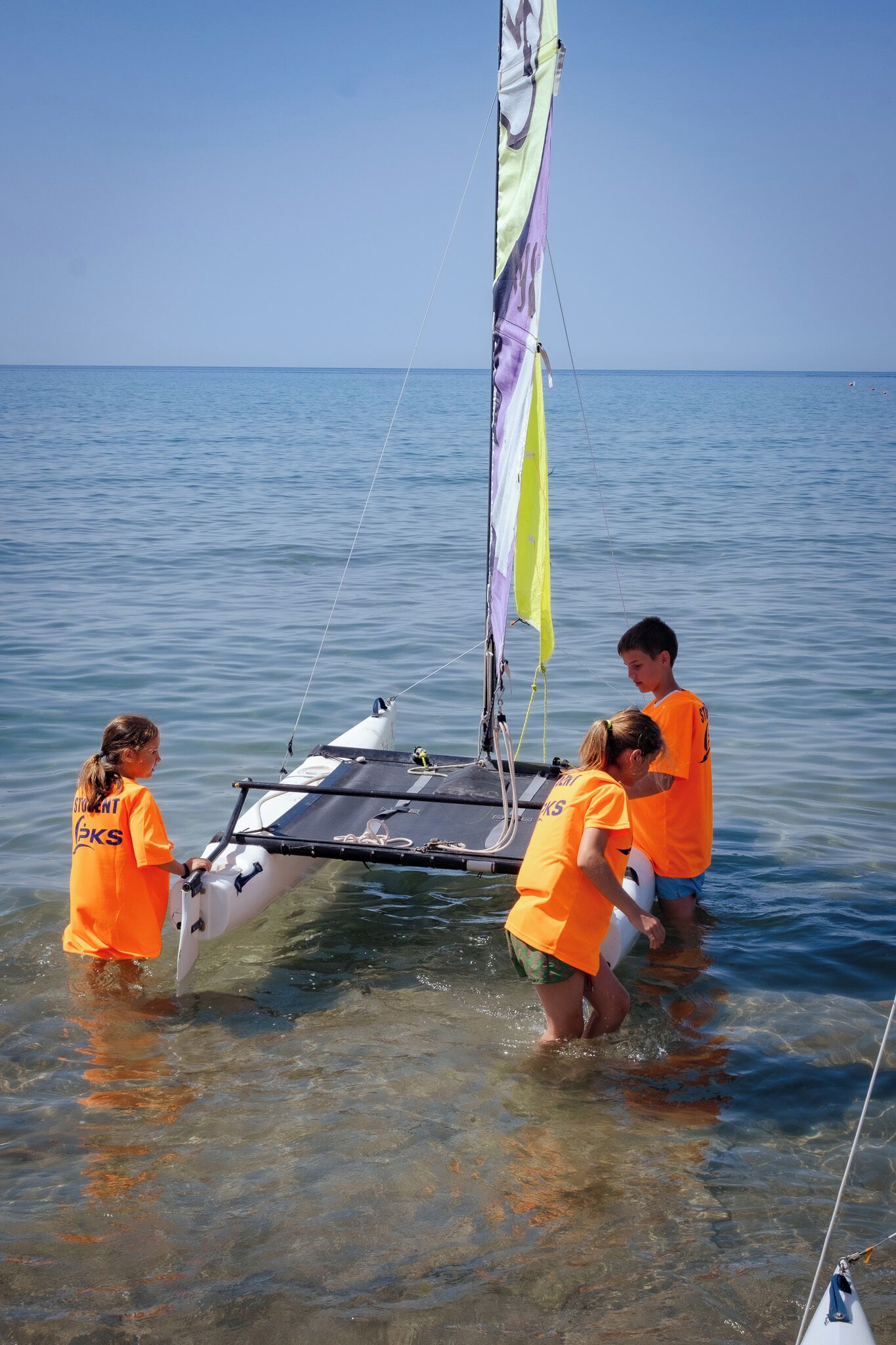 A small sailboat with a sail, being handled by people in orange shirts.