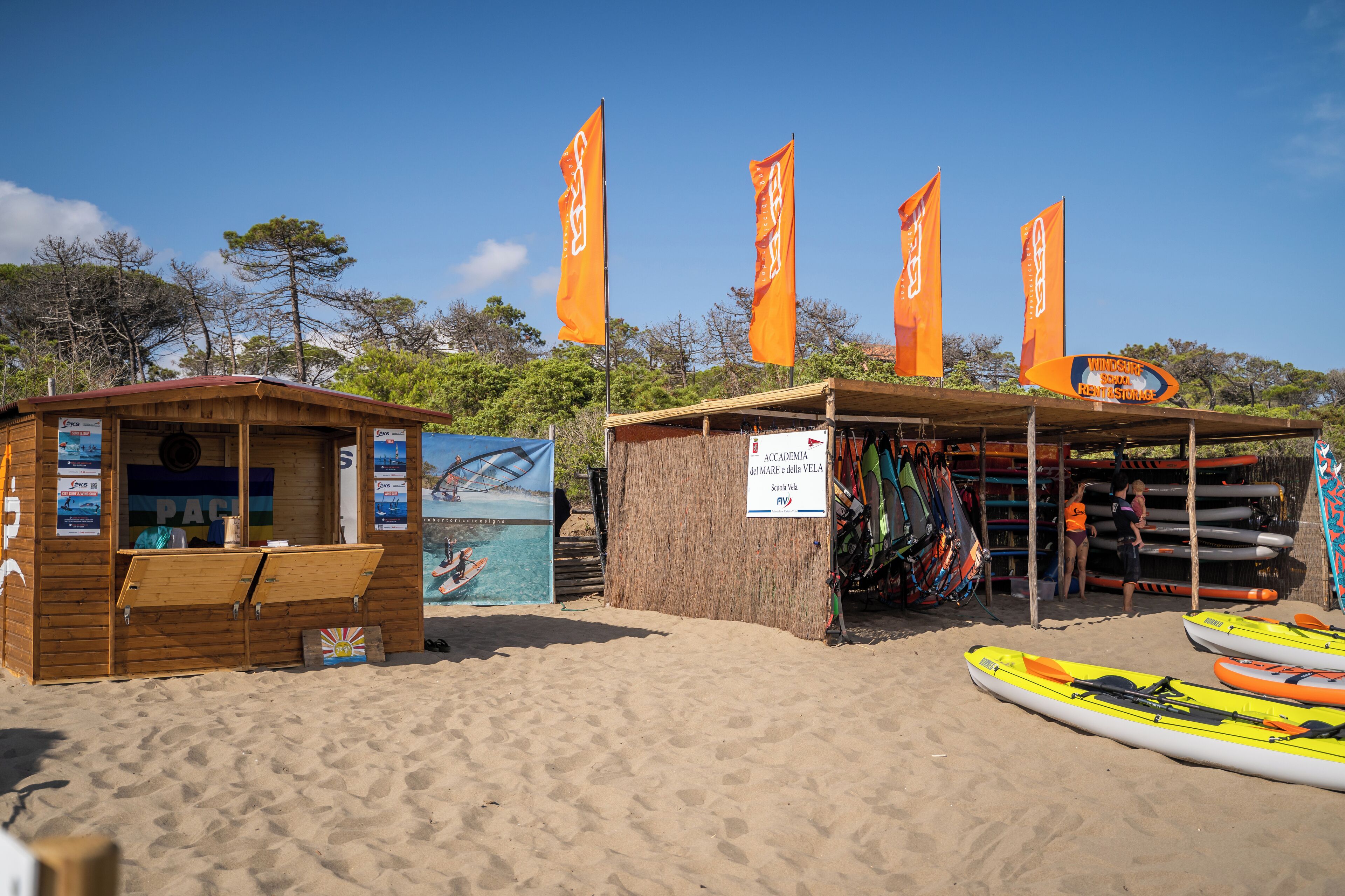A beachside building with a wooden structure, a sign for 'Accademia del Mare e della Vela', and several kayaks and canoes.
