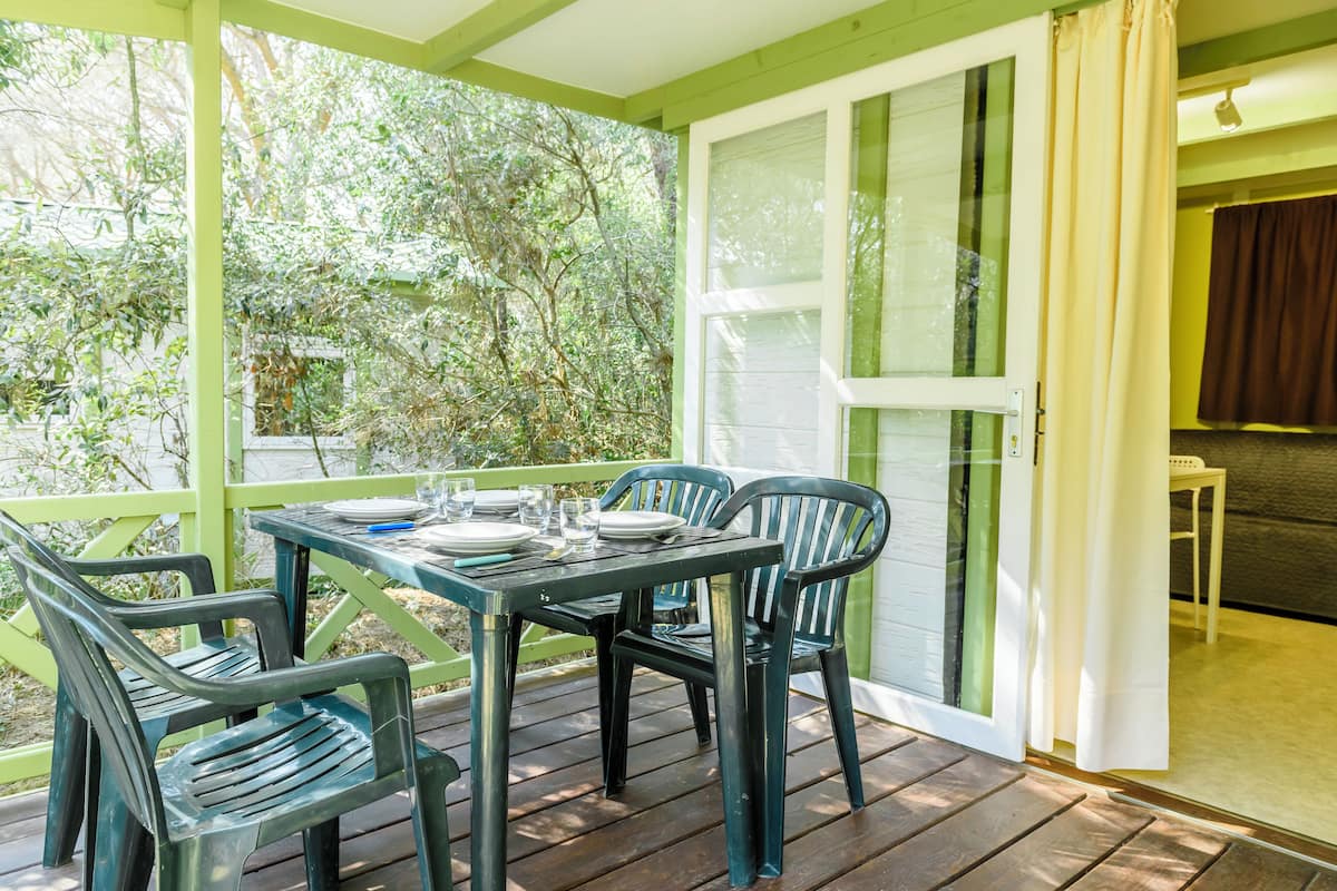 A covered patio with a table set for four, surrounded by green chairs, and a view of trees through the glass door.