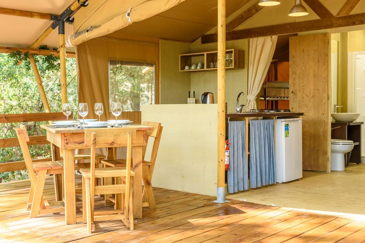 A wooden deck with a table and chairs, a kitchen area with a sink and stove, and a view of greenery outside.