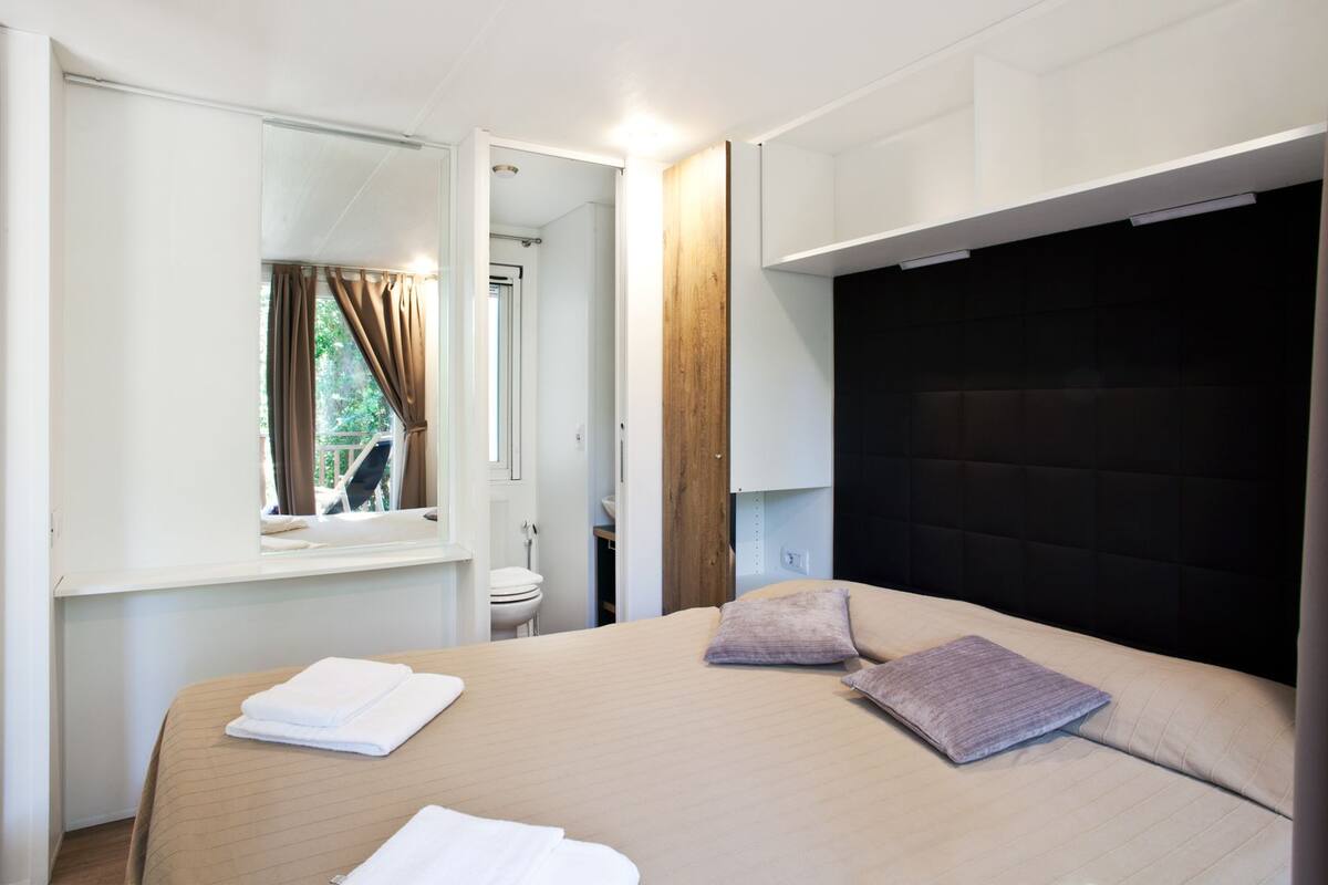 A bedroom with a bed, a window, a mirror, and a view of the outdoors.
