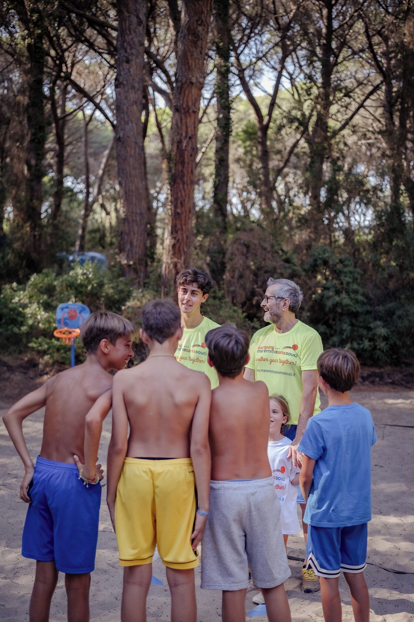 Un groupe de personnes se tient à l’extérieur, certaines torse nu, dans un environnement naturel avec des arbres et un panier de basket en arrière-plan.